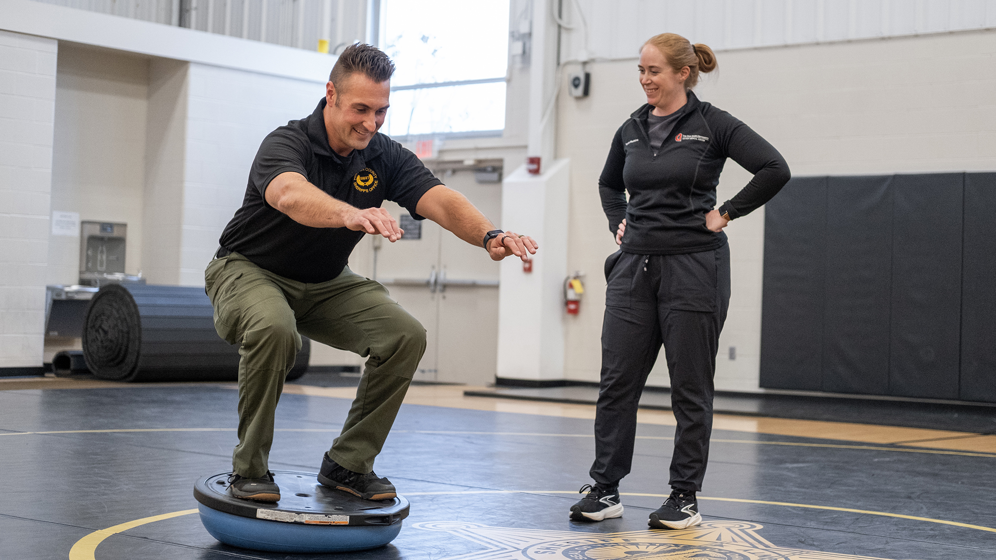 A sheriff's officer balances on a platform as an athletic trainer monitors his performance