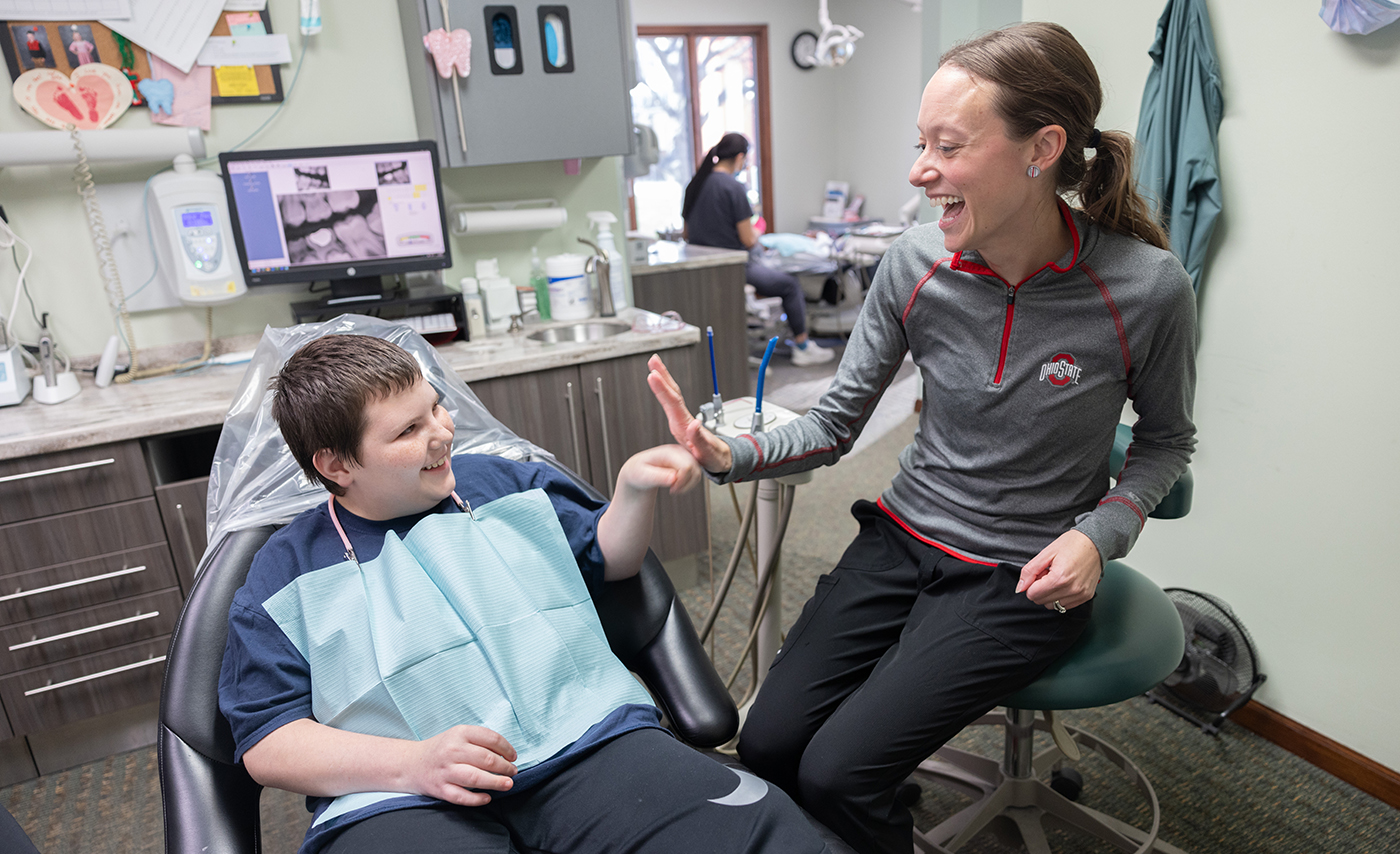 A dentist gives a high five to a young male patient who is reclining in a dentist's chair.