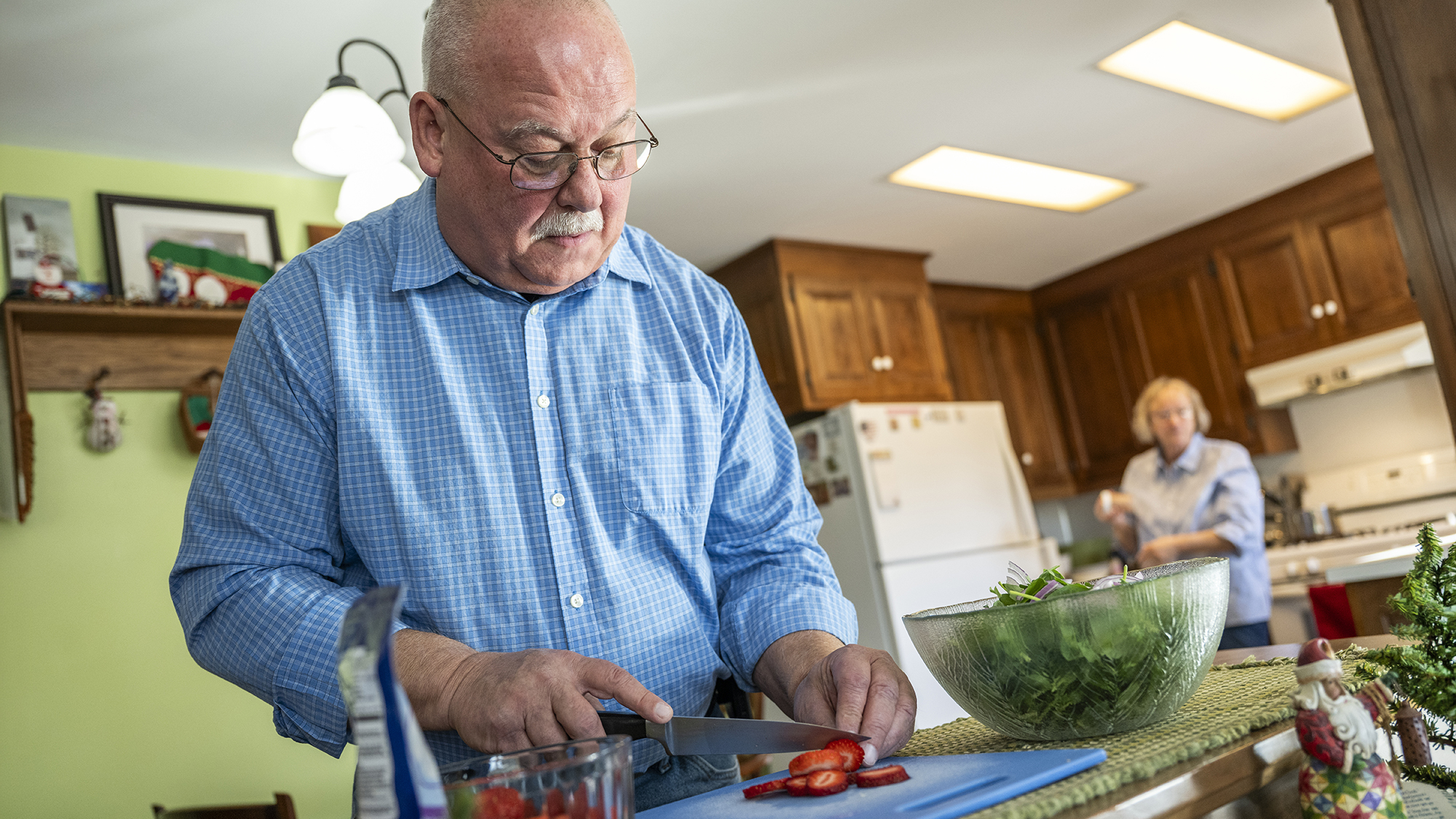 A man in a blue shirt chops strawberries in a kitchen as his spouse looks on in the background.