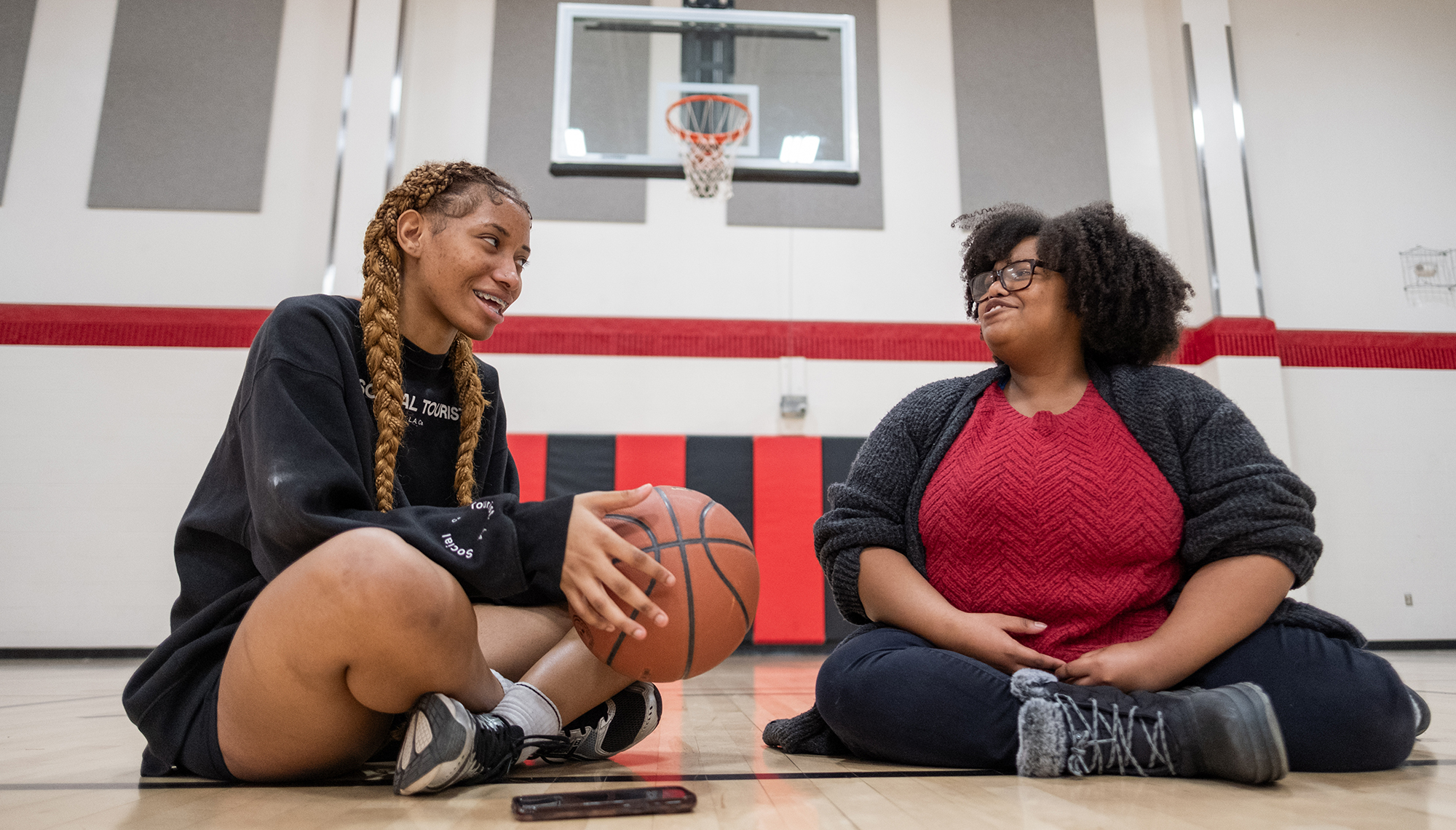 Two young people sit cross-legged next to each other on a basketball court with the basket behind them