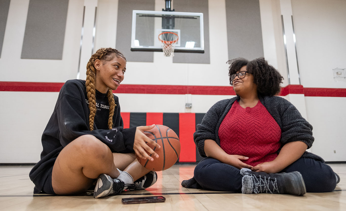Two woman, the one on the left holding a basketball, talk while seated cross-legged on a basketball court