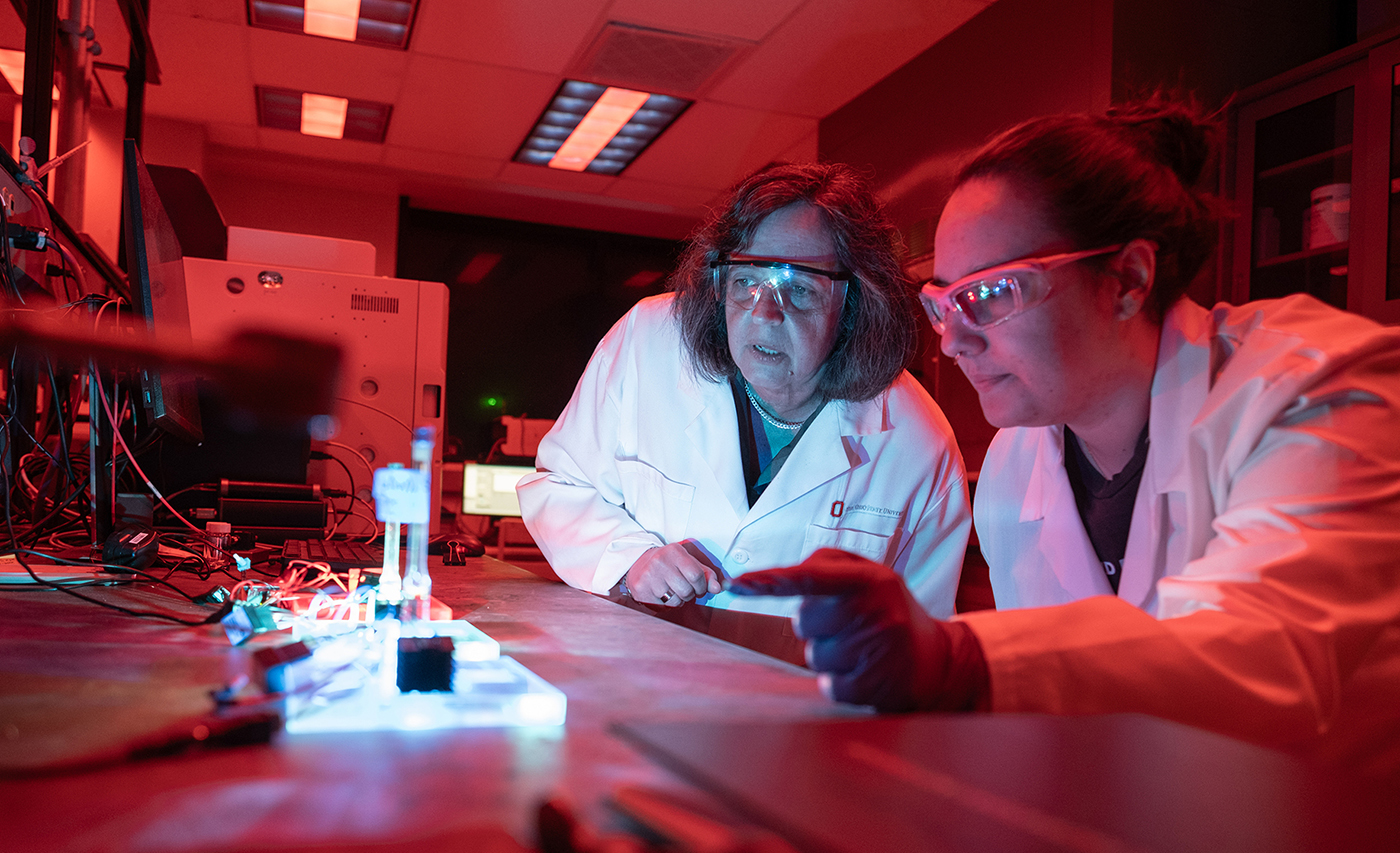 Two researchers wearing protective eyewear examine an experiment in a lab that is lit with a scarlet light