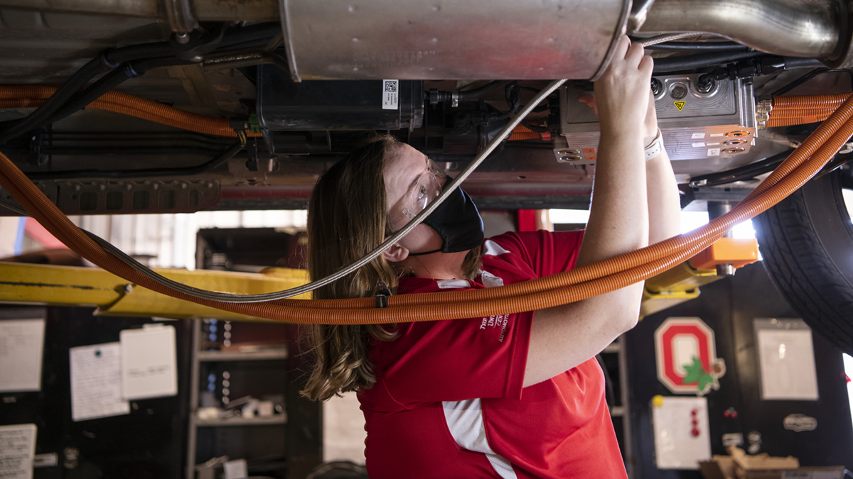 A researcher wearing a mask works on the undercarriage of an automobile.
