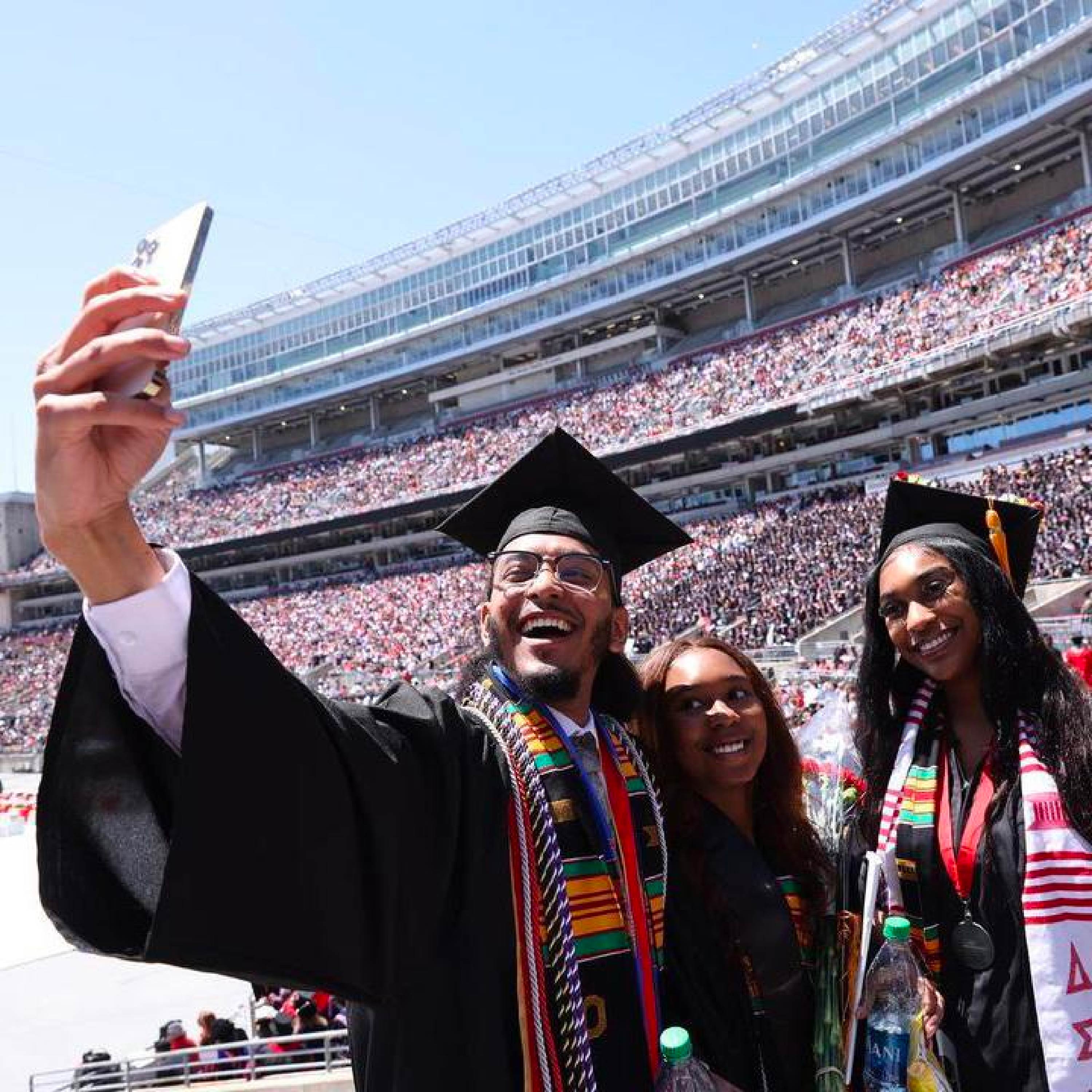 A student graduating from Ohio State in Ohio Stadium smiles while taking a selfie of himself and two friends.