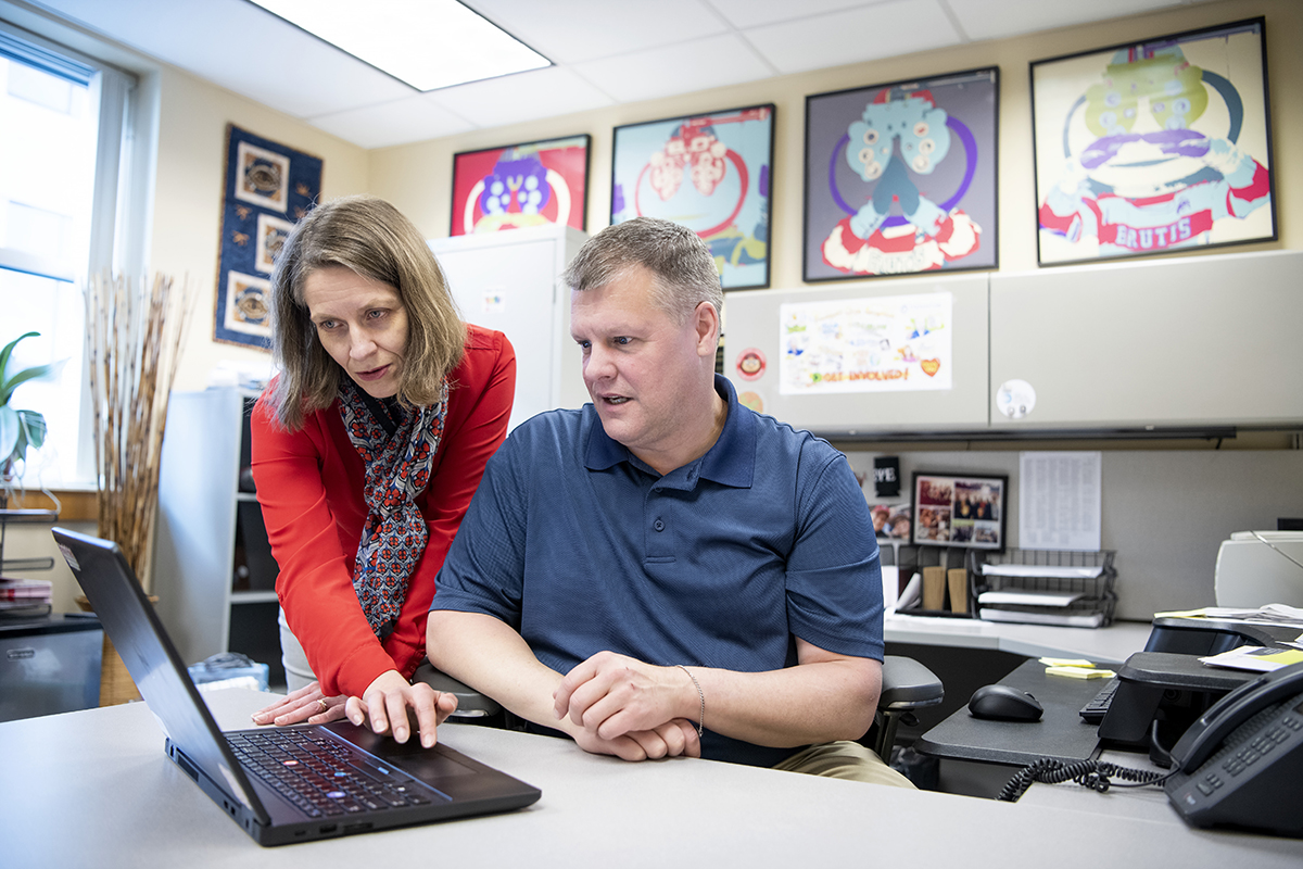 A woman stands next to a seated man while both look at an open laptop computer