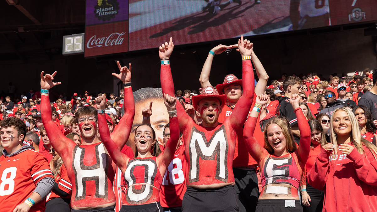 Students in red body paint with the word "HOME" spelled out on their chests celebrate at an Ohio State football game
