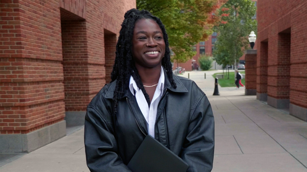 Rachel Boayke looking off camera and smiling as she stands in-between two buildings on campus with her gap, gown and diploma