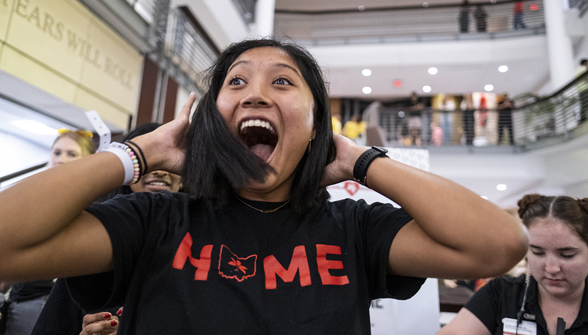 A student in the Union smiling with their mouth open and hands on their heads wearing a shirt that says 