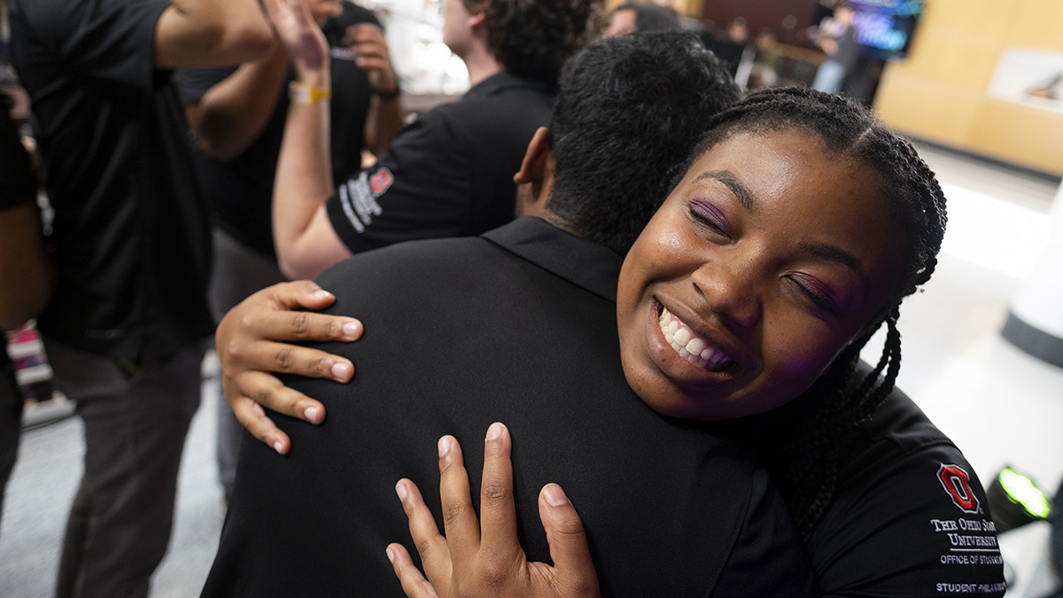 A close up of two students embracing each other in a hug. One of the students is smiling with their eyes closed while facing the camera