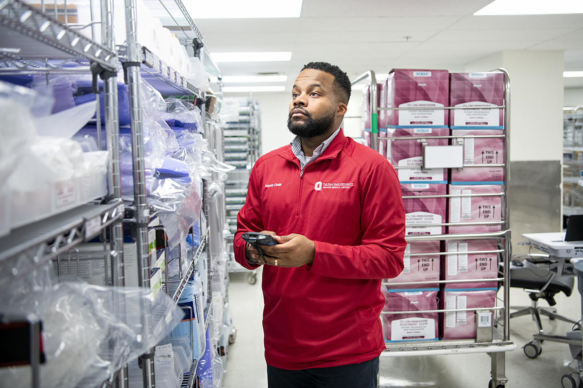 A man in a red pullover holds a device in his hand while looking at a rack of medical supplies.