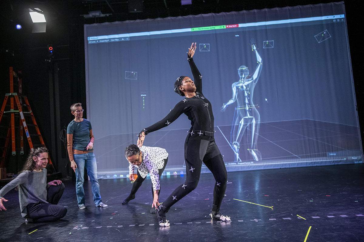 A dancer in a motion capture suit performs movements that are displayed on a digital screen behind her as three women faculty members look on.