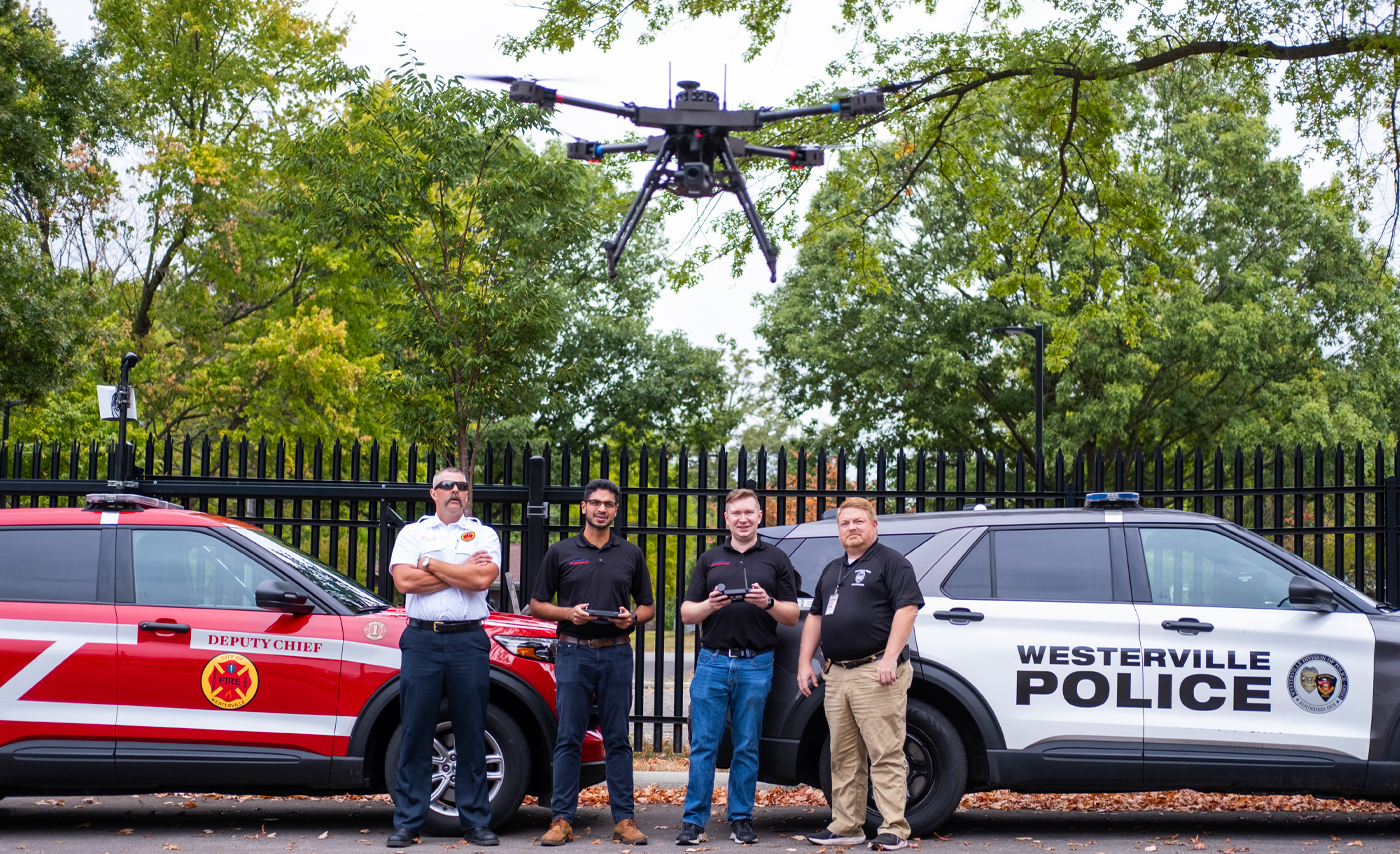 A drone flies in front of two students and two first responders.