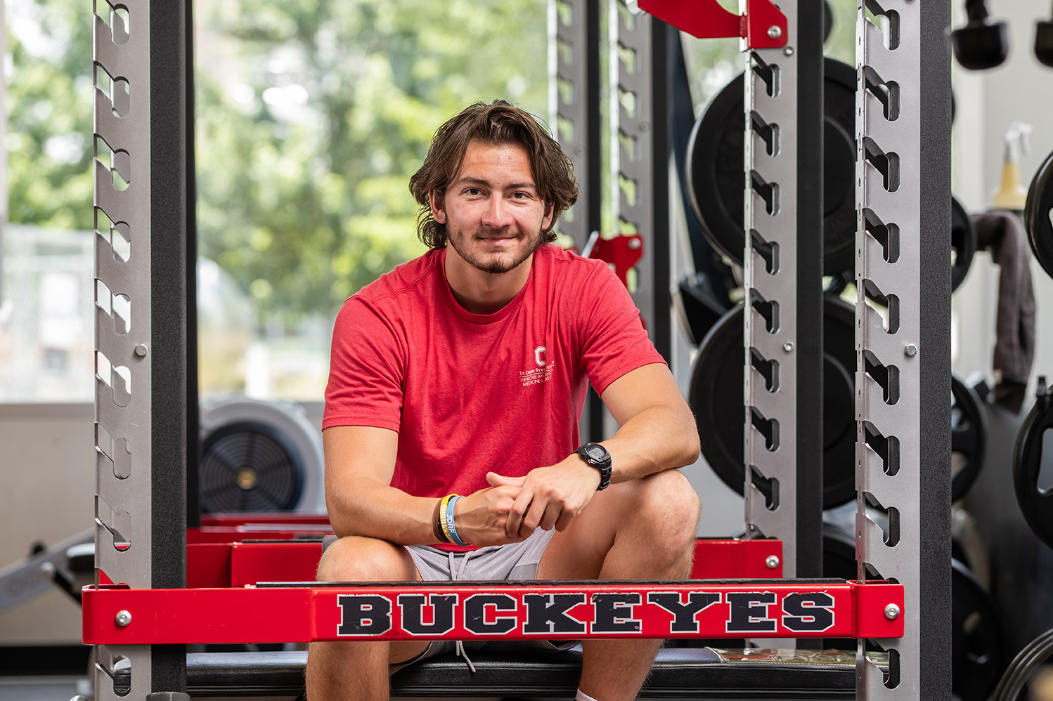 A young man sits in a weight room in front of a bench that reads "Buckeyes"