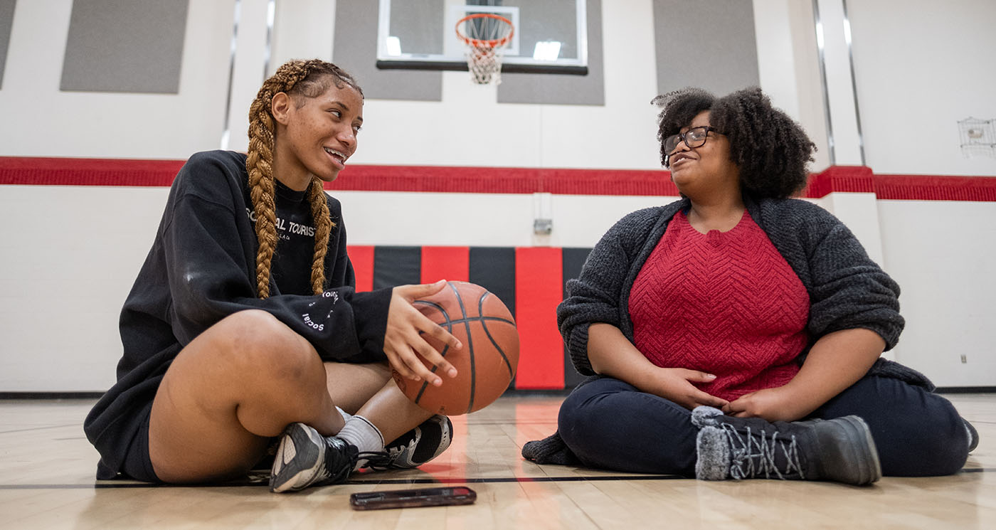 Two women sit on a basketball court and have a conversation