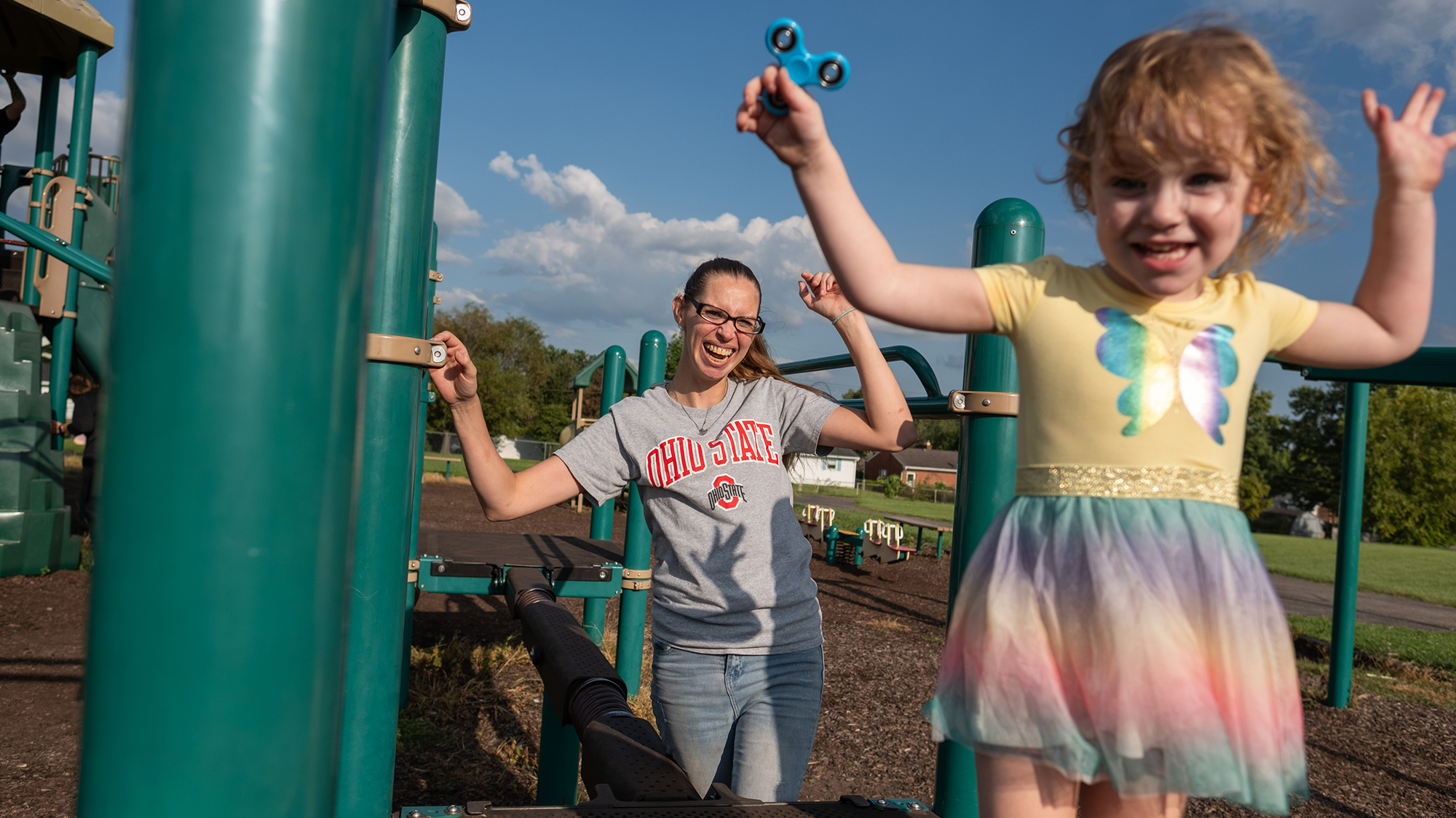 A young girl raises her hands while playing on playground equipment while her mother smiles behind her