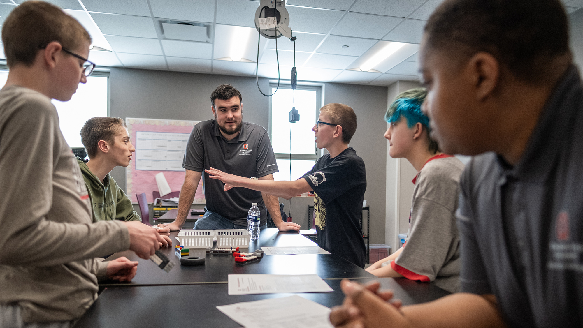 A man leans on a table in a classroom and looks on intently as students discuss a project