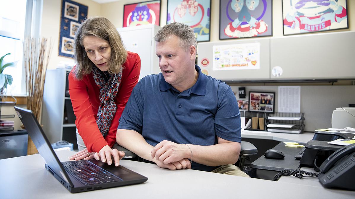 A woman stands next to a seated man while both look at an open laptop computer. 