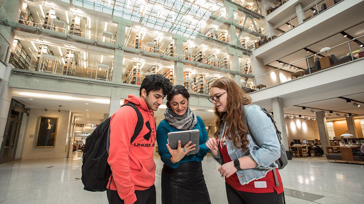 Three students stand together and look at a tablet computer.