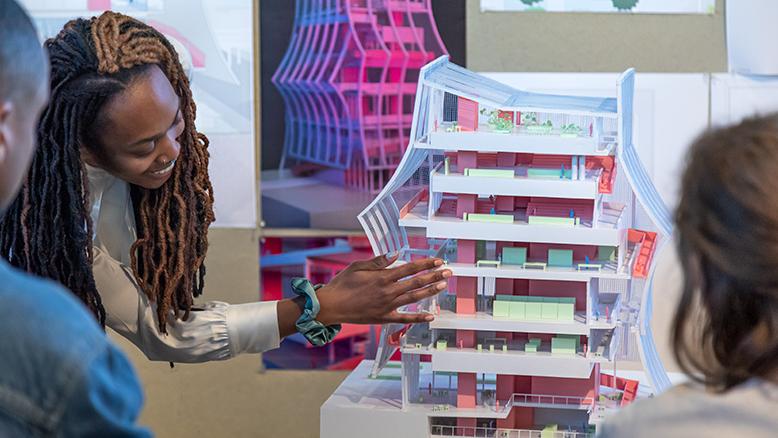 A student gestures at a building model in pink and green showing interior volumes. In the background are renderings of the design in black, blue and pink.