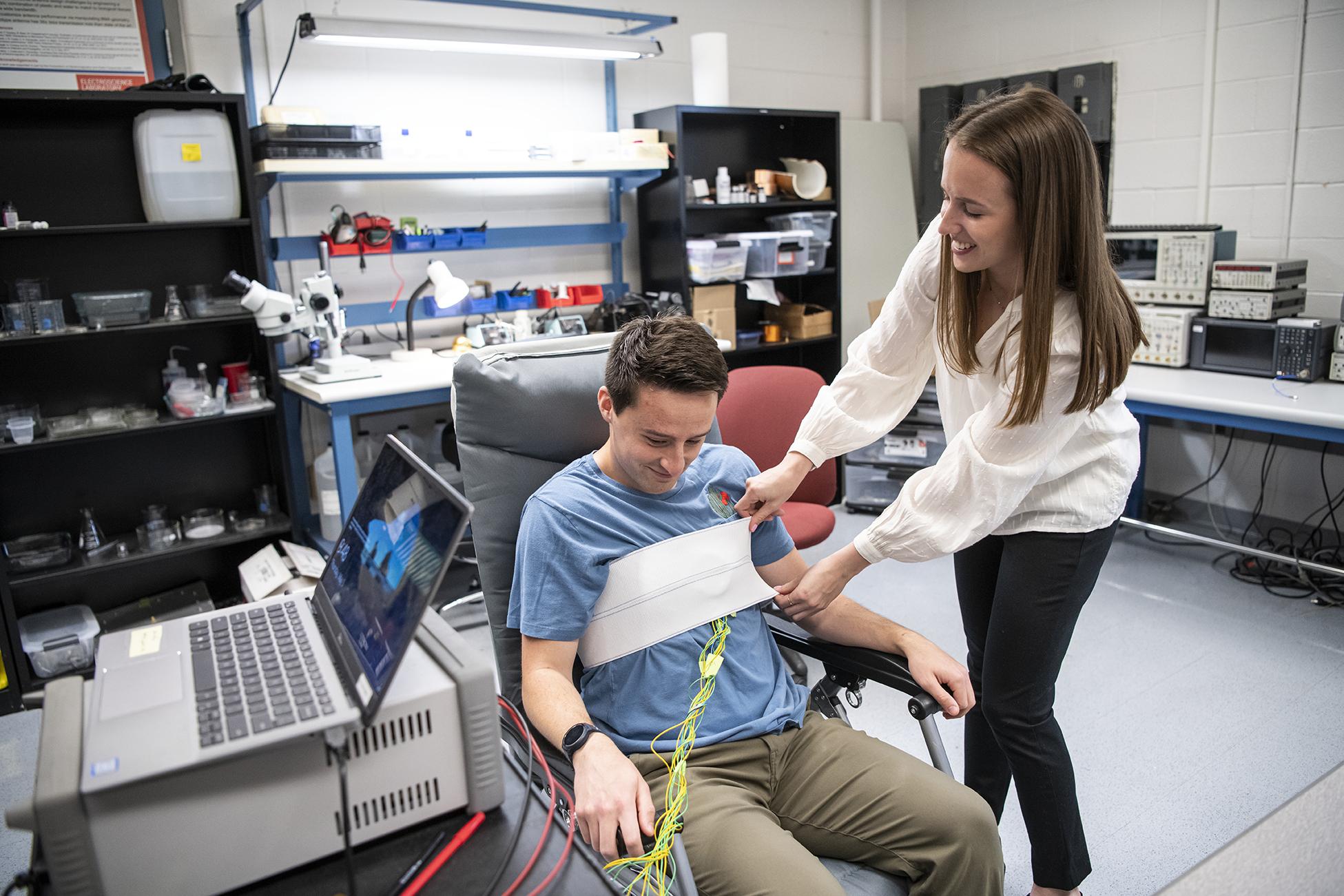 A young woman applies sensors to a man seated in a chair in a lab environment