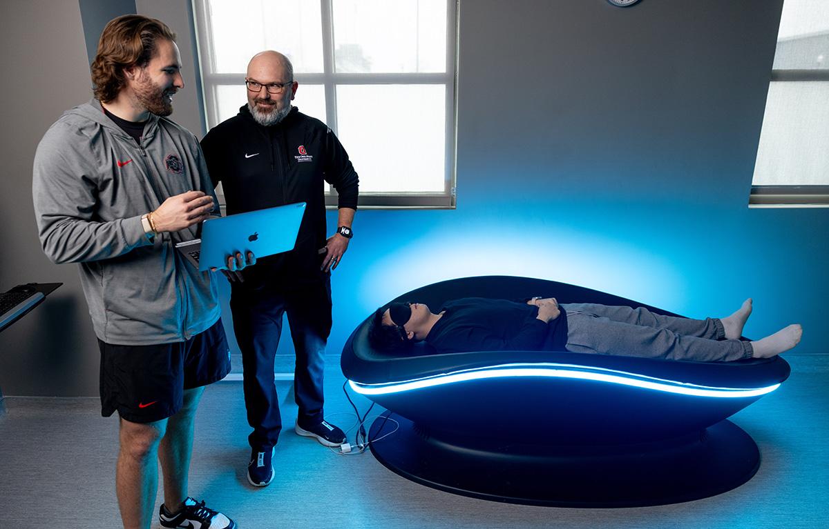 Two men stand next do a machine that has a masked person laying on it.