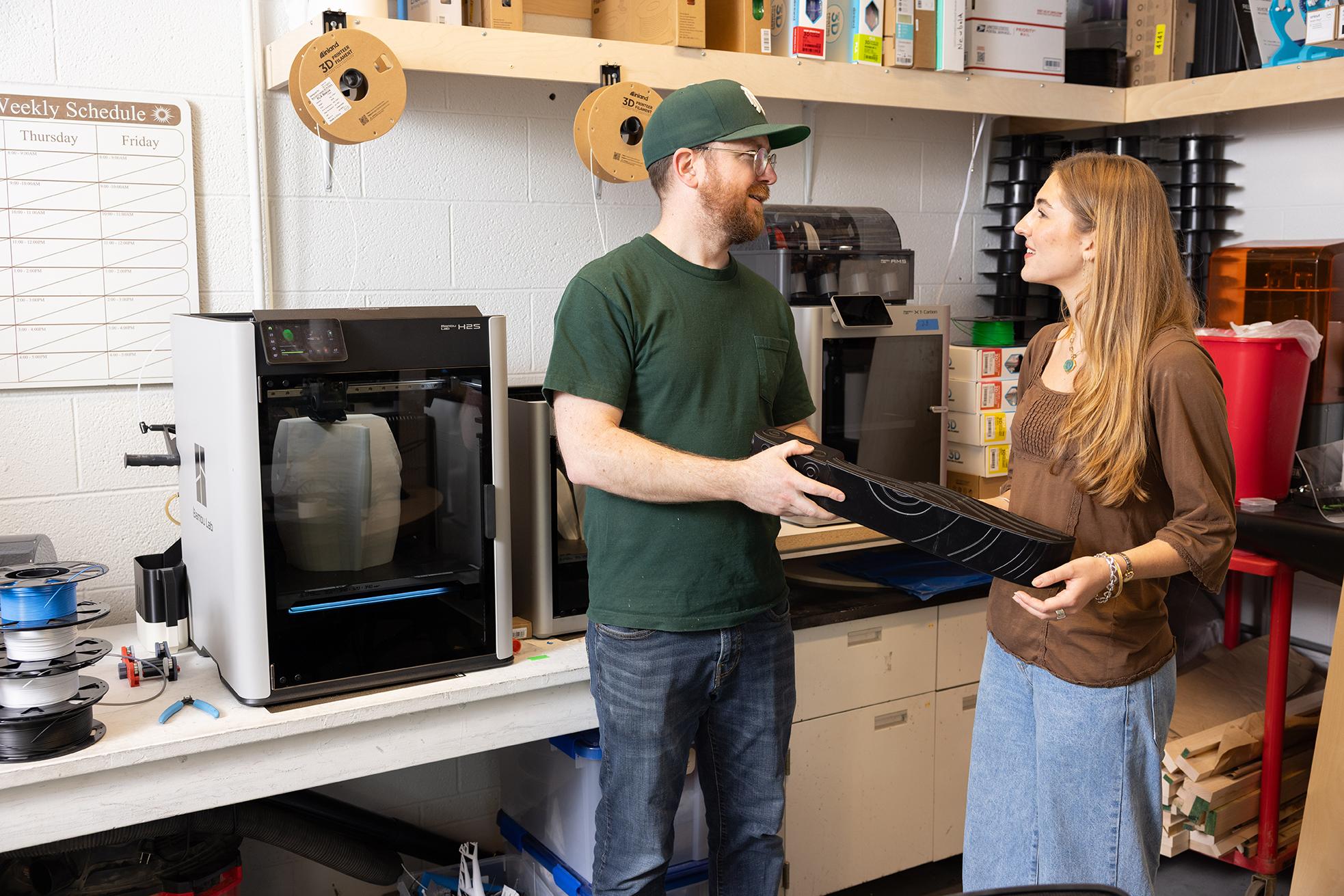 A young man and woman talk together in a lab with a 3D printer to their left