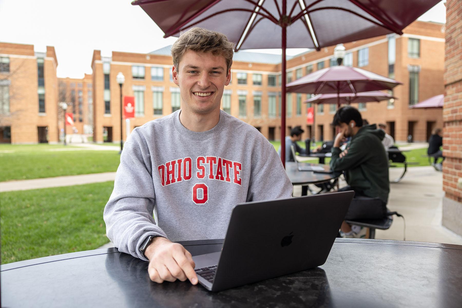 A portrait of Ohio State student Reed Kroger sitting outside on the Fisher College of Business campus with his laptop open in front of him