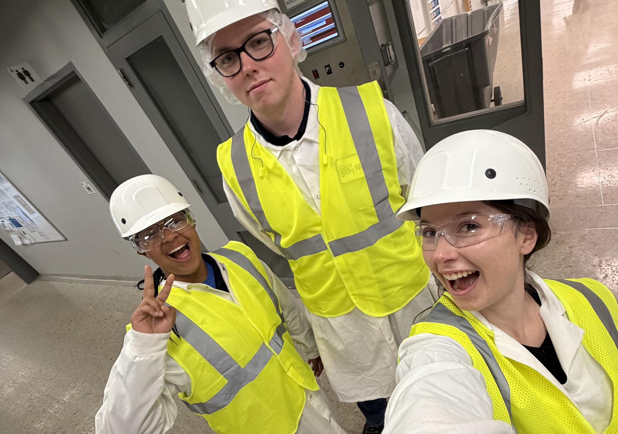 Three young people in hardhats, protective eyewear and yellow safety vests stand outside a lab